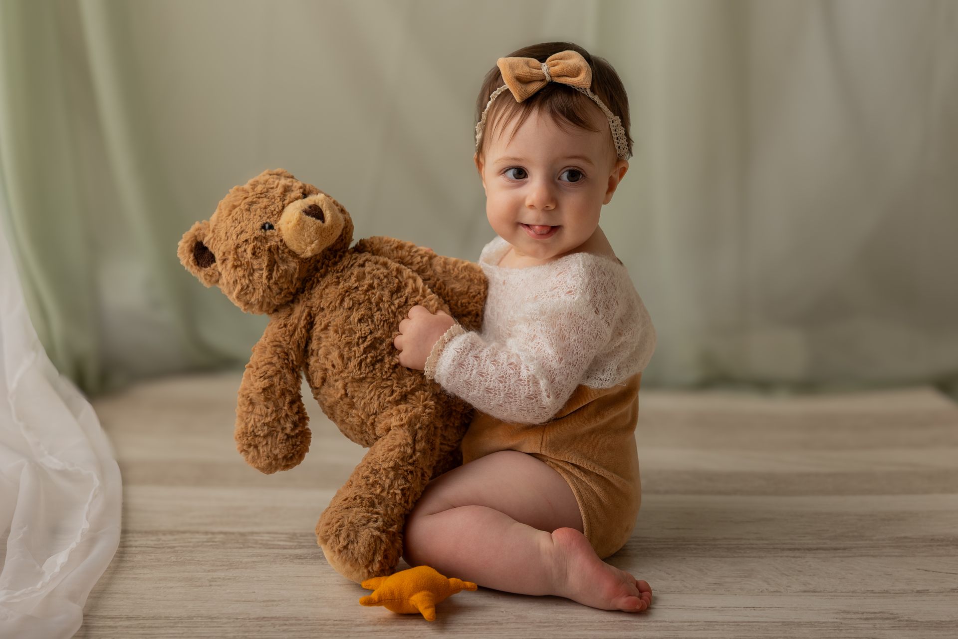 Seduta sul parquet chiaro, la bambina abbraccia un grande orsetto marrone davanti a una tenda verde