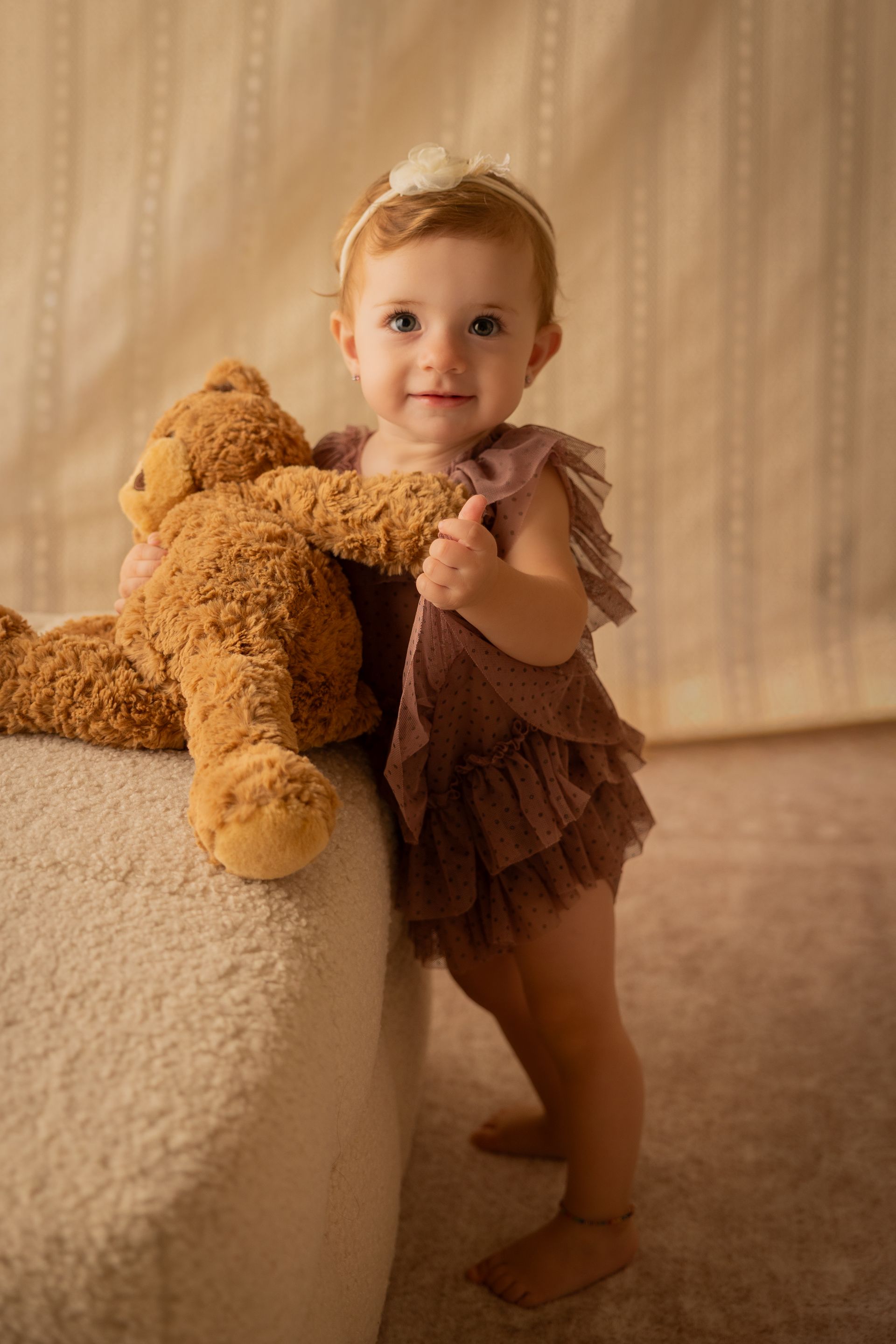 Bambina in piedi appoggiata a un pouf bianco con orsacchiotto, vestitino a balze color prugna