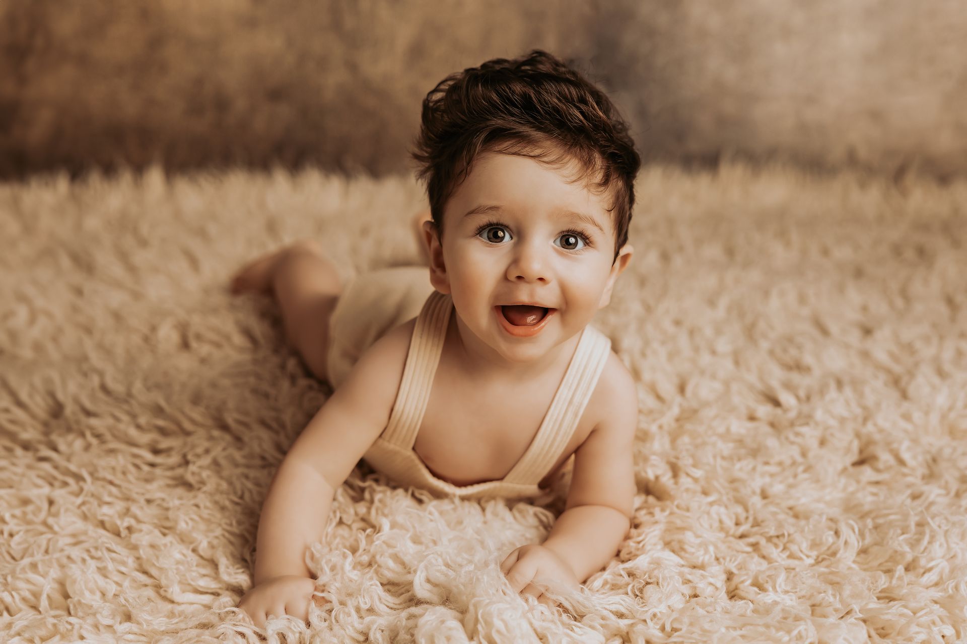 Bambino a pancia in giù su tappeto peloso beige, sorride a bocca aperta guardando in alto