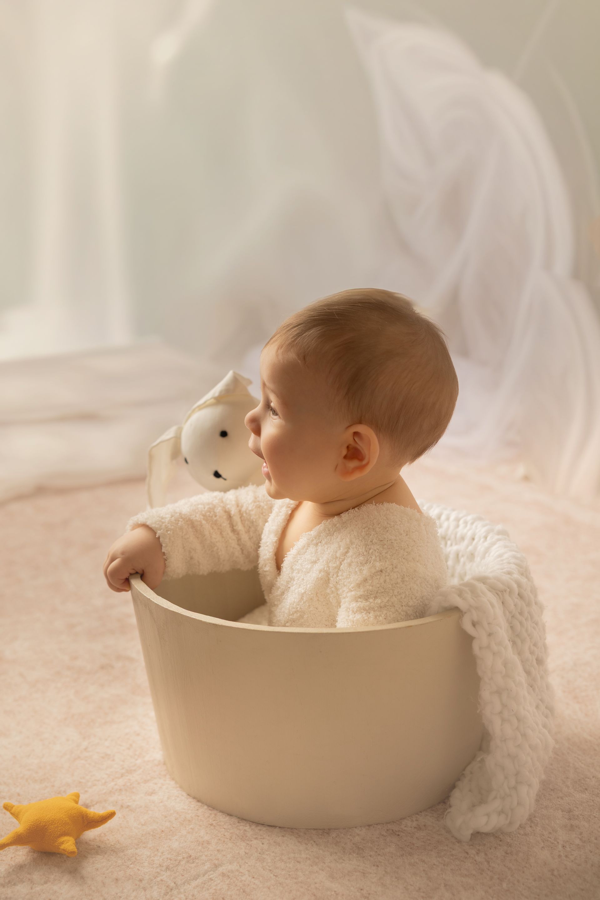 Di profilo dentro un mastello chiaro, il bambino guarda un orsetto bianco fra tende luminose
