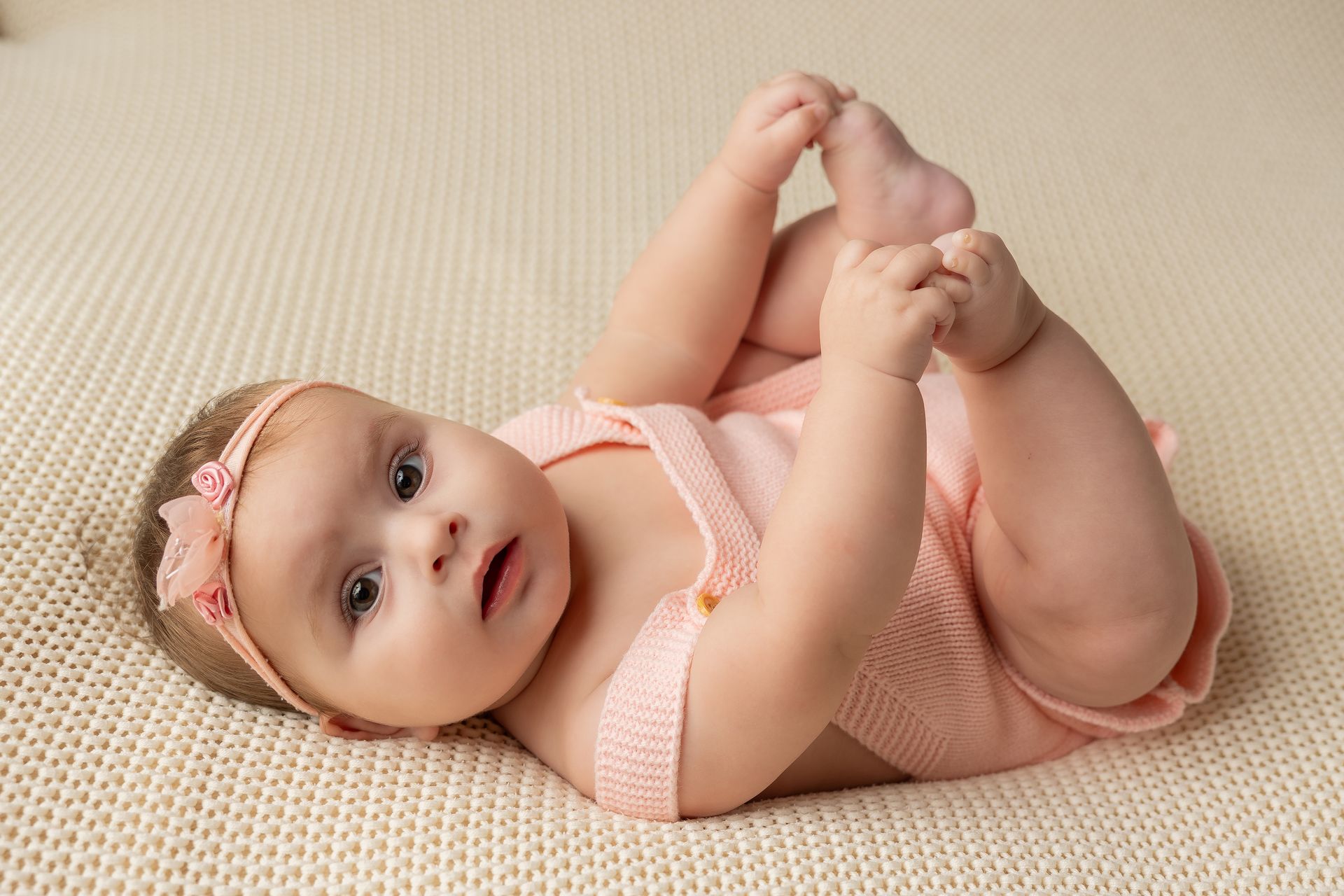 Bebè su un fianco che si tiene i piedini con le mani, in body rosa su una coperta panna a maglia