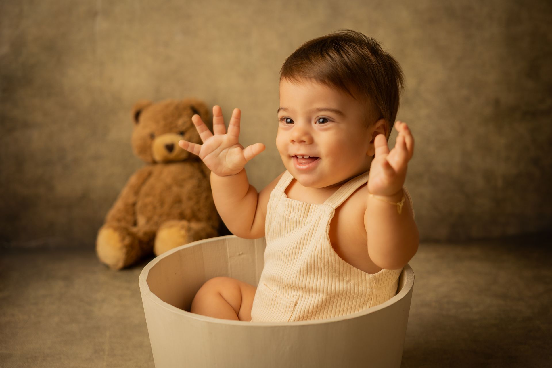 Bambino in tutina panna alza le mani aperte dal mastello, un orsetto sfocato dietro di lui
