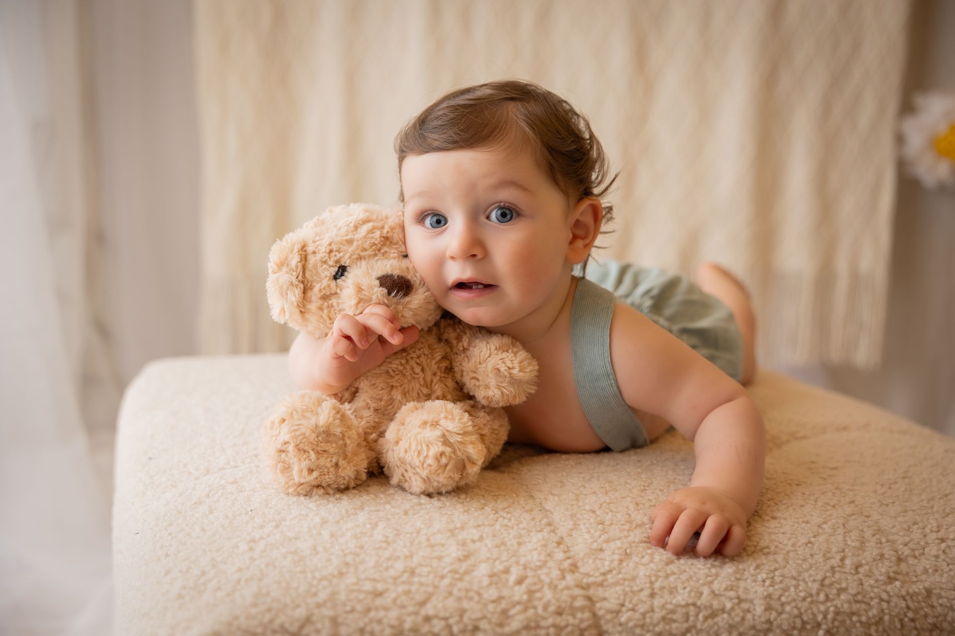 A pancia in giù su un pouf color panna, il bambino stringe un orsetto beige