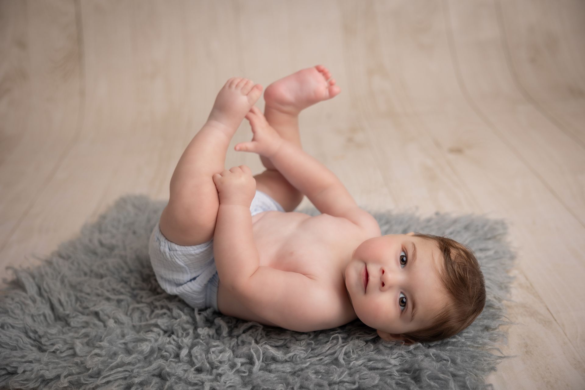Bambino disteso su un tappeto grigio a pelo lungo si afferra i piedini e guarda in camera