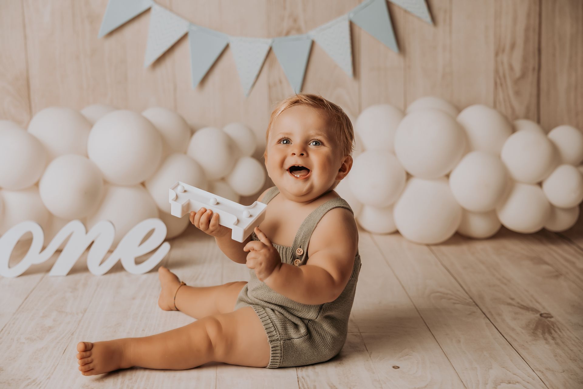 Bimbo sorridente seduto tra palloncini bianchi con scritta ONE in legno e bandierine azzurre