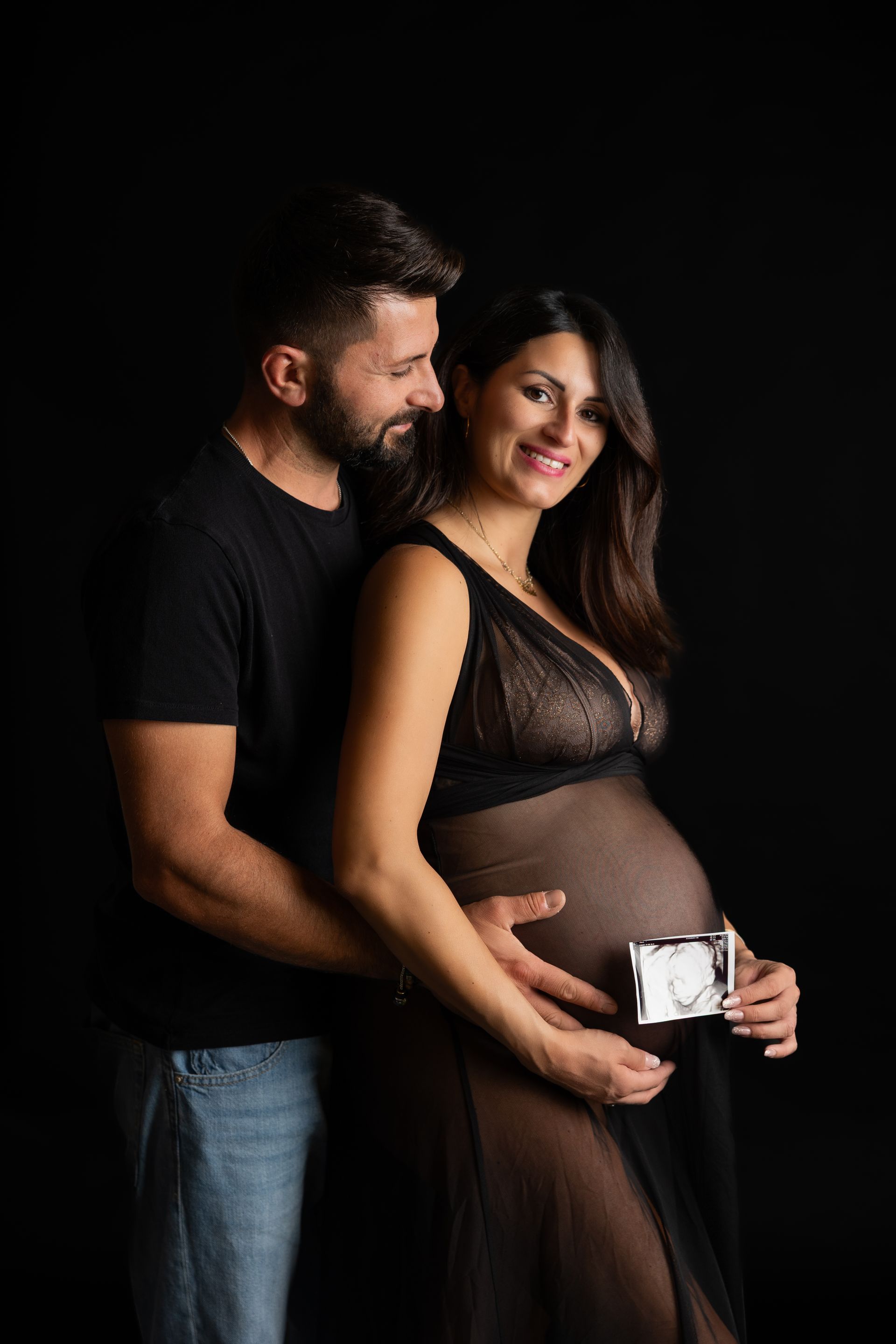 Lei in abito nero trasparente regge l'ecografia sorridendo, lui la abbraccia da dietro su fondo nero