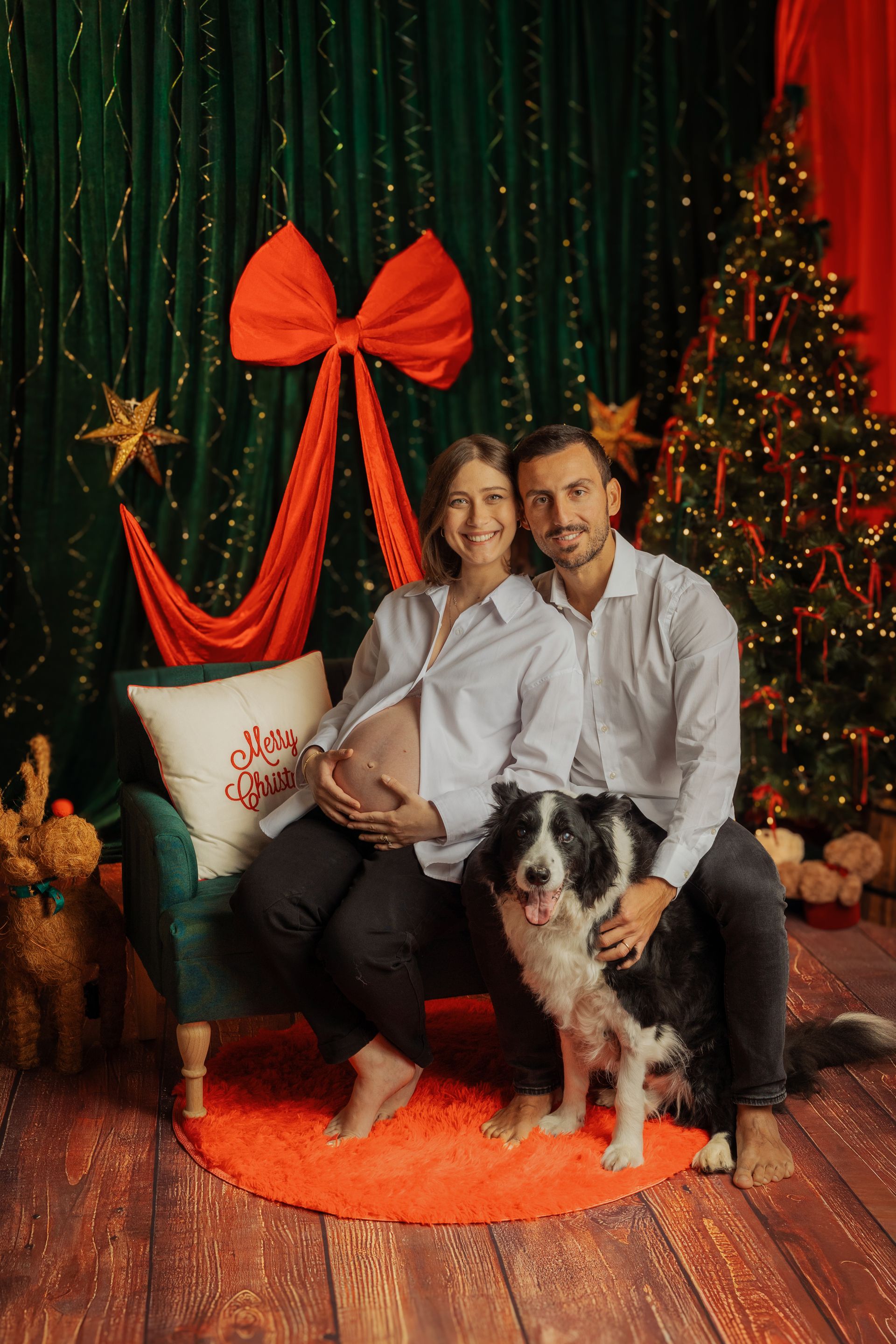 Una coppia seduta sulla poltrona verde con il border collie, fra l'albero illuminato e il fiocco rosso