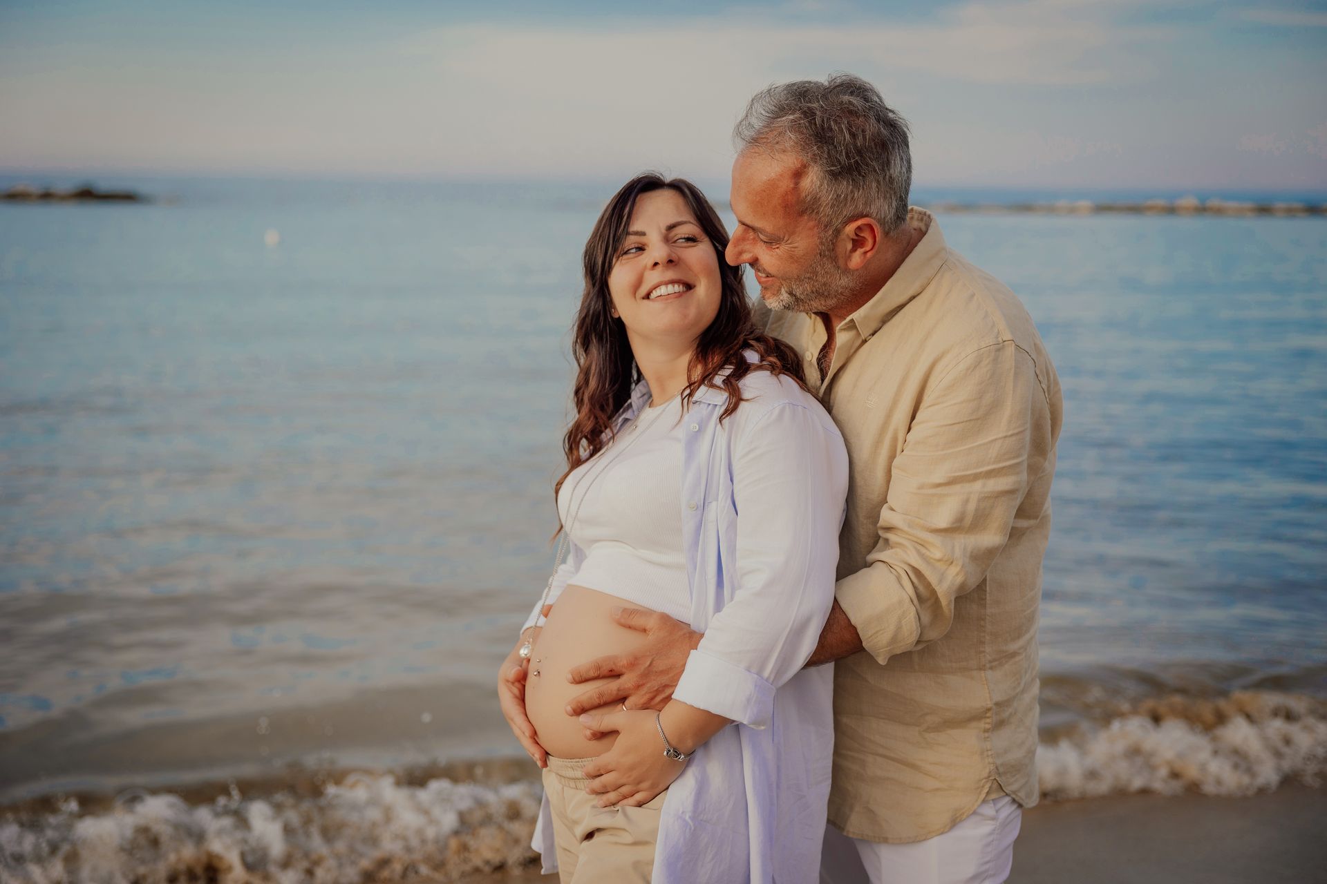 Coppia che ride sulla riva, lei con camicia aperta e pancione scoperto davanti al mare azzurro