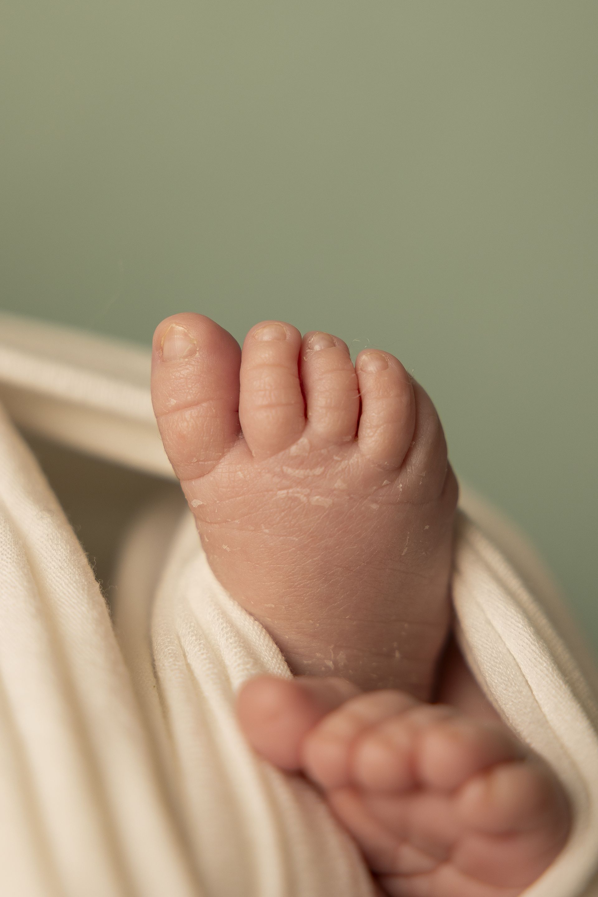 Dettaglio di un piedino minuscolo poggiato su telo crema con fondo verde salvia sfocato