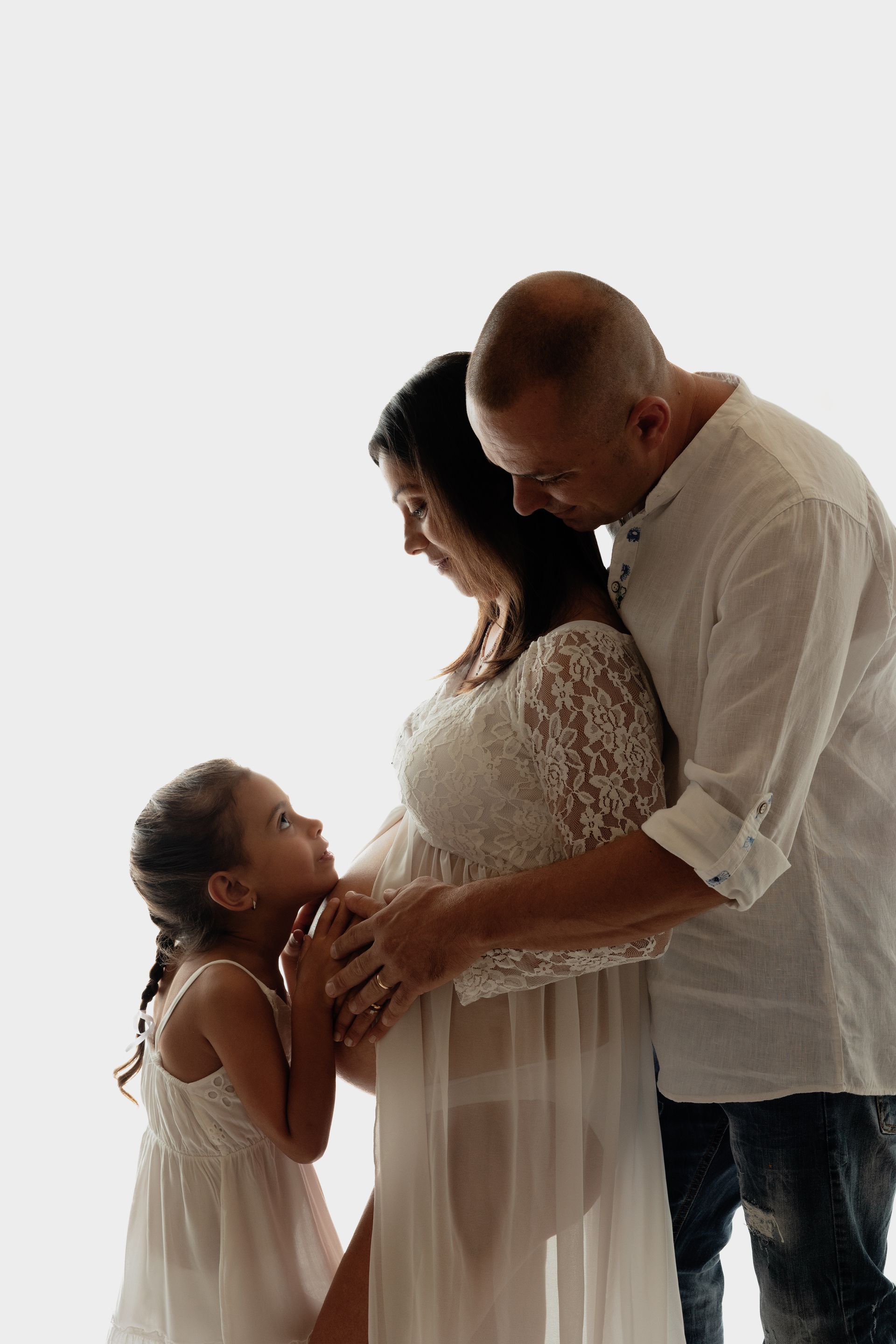 Bambina in abito bianco guarda in alto toccando il pancione della mamma abbracciata dal papà