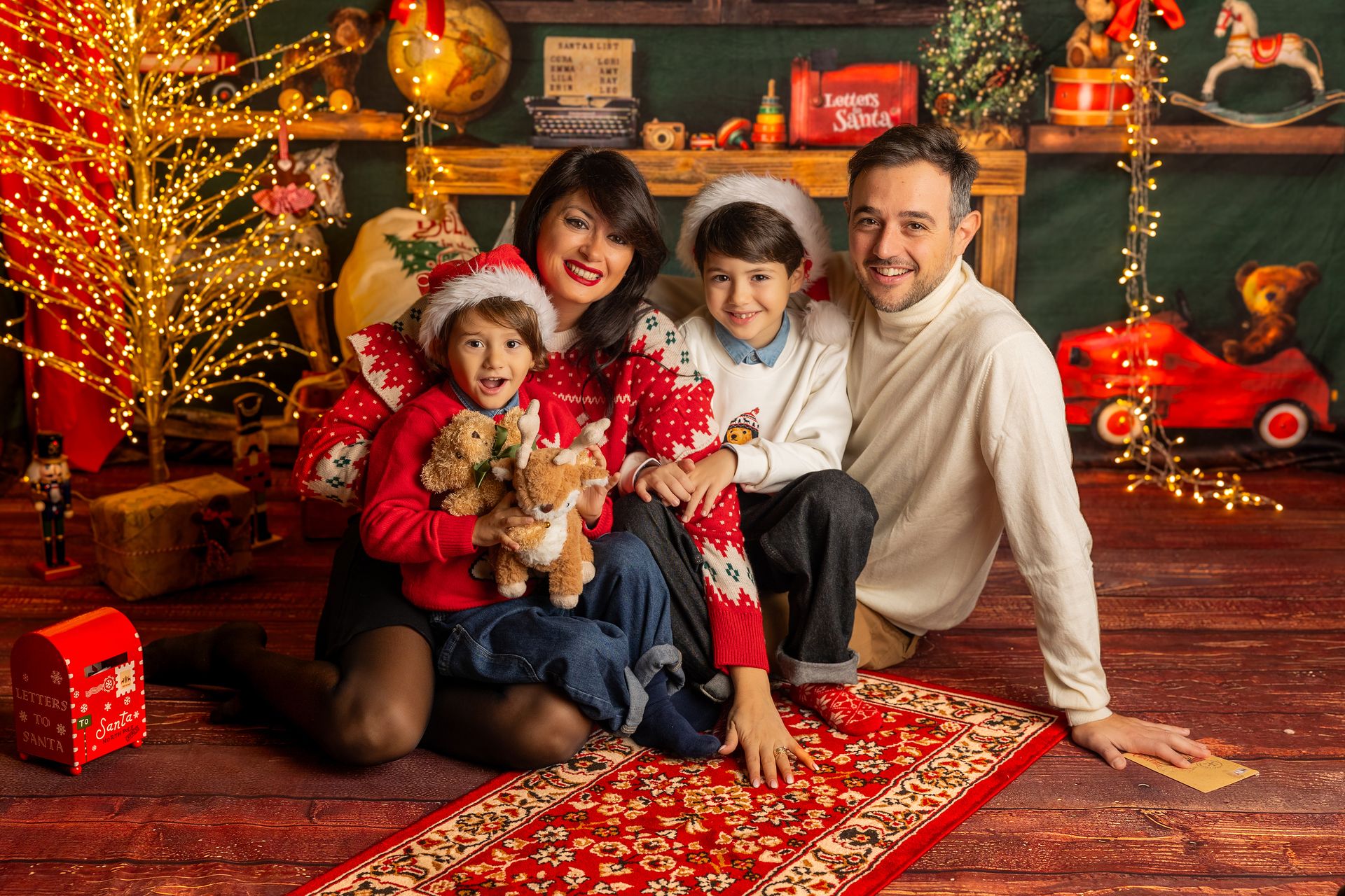Famiglia di quattro seduta sul tappeto rosso del set natalizio, fra gli alberi illuminati e i giocattoli di legno