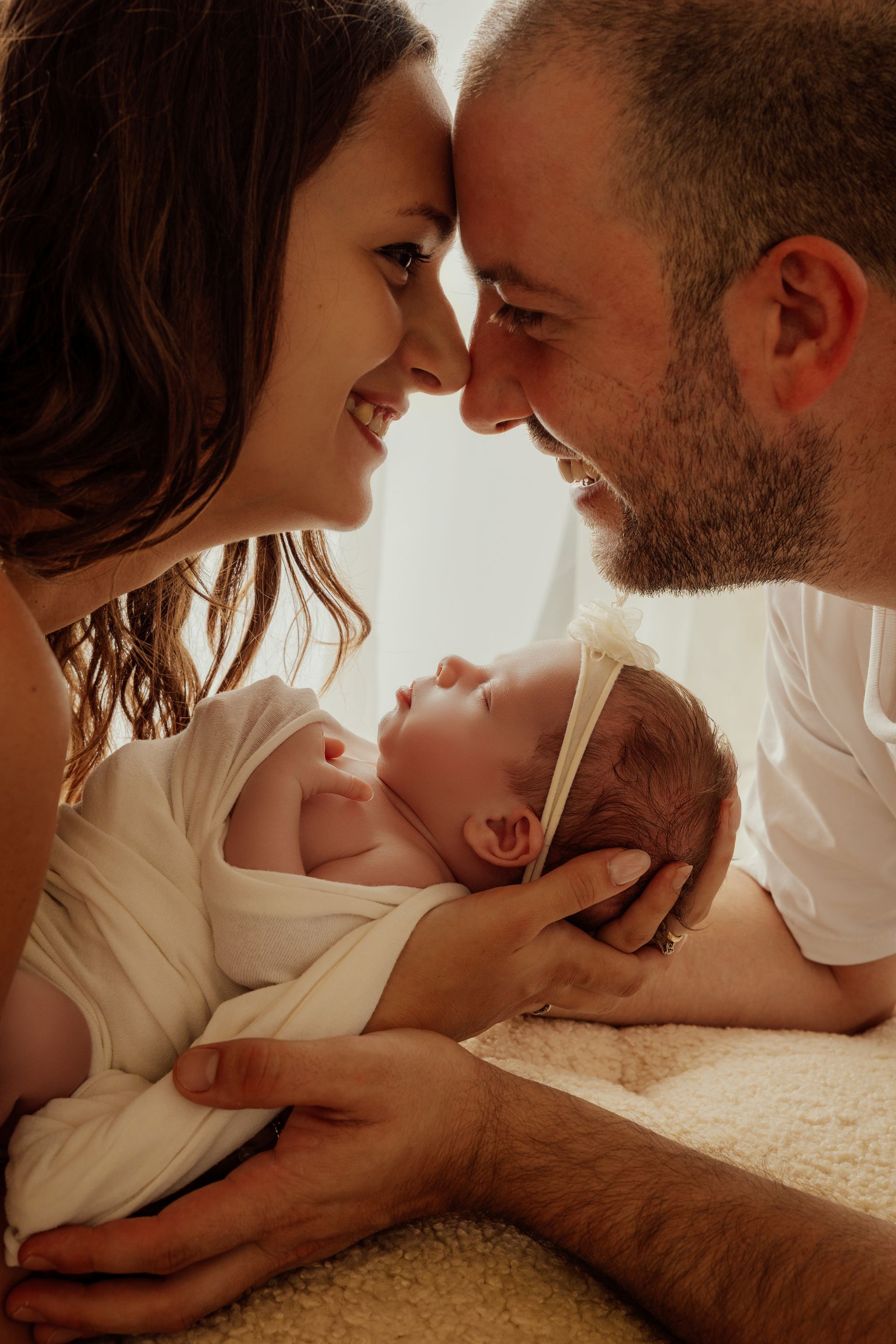 Genitori sorridenti fronte contro fronte mentre tengono fra le braccia la neonata avvolta nel bianco