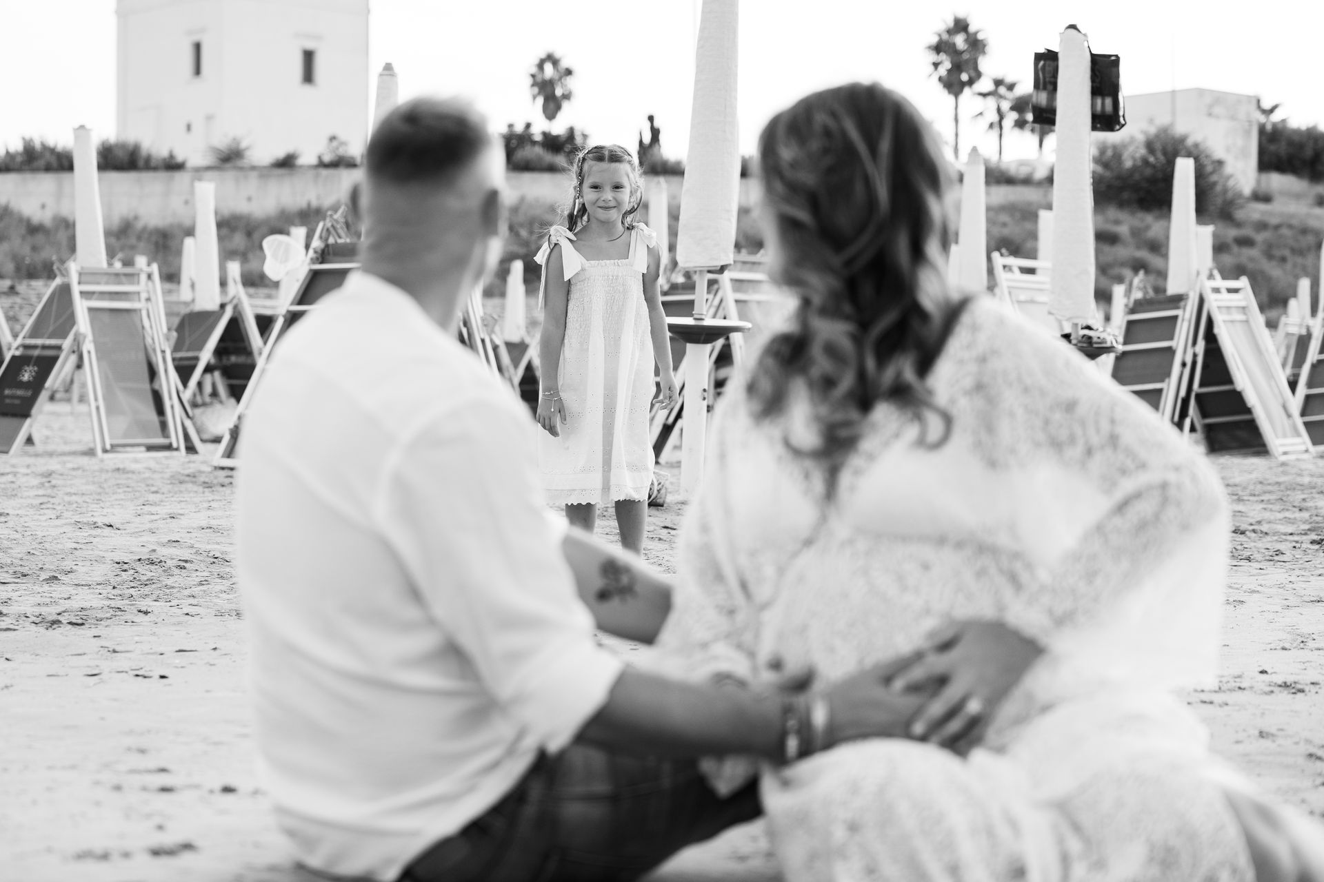 Genitori seduti sulla sabbia guardano la figlia in piedi davanti a loro tra gli ombrelloni, scatto in bianco e nero