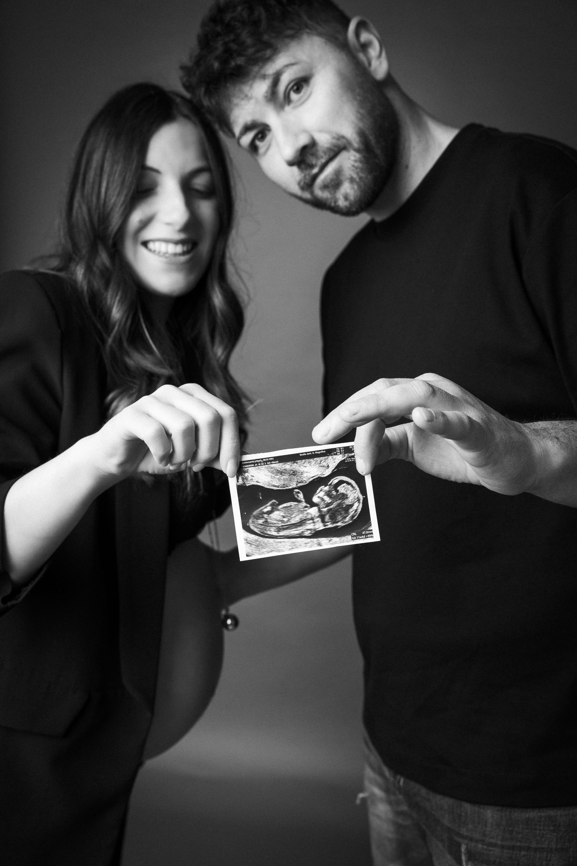 Due futuri genitori mostrano alla camera l'ecografia del bambino, teste appoggiate e sorriso complice in monocromia