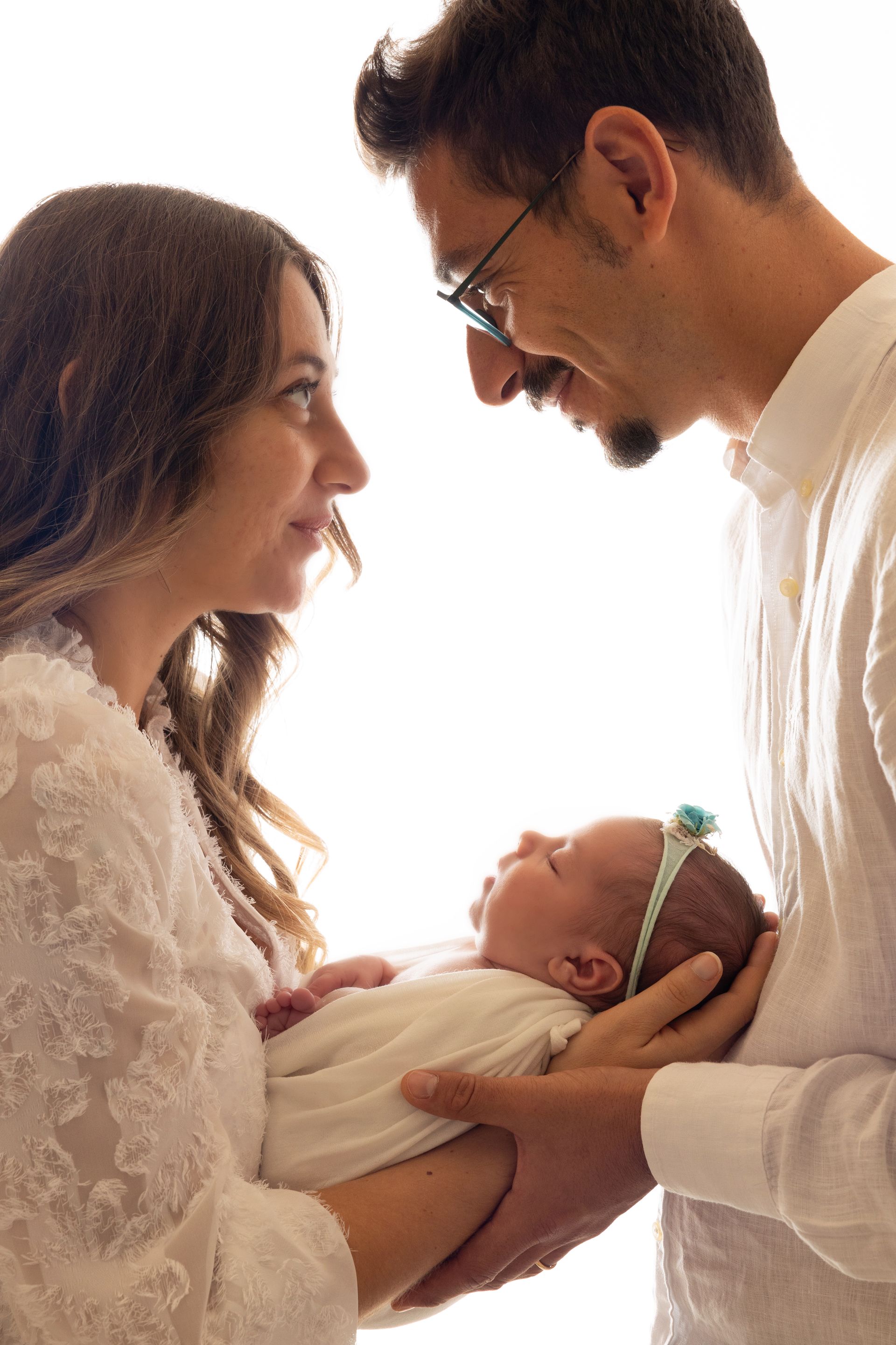 Genitori che si guardano negli occhi tenendo la neonata fasciata, luce diffusa e abito di pizzo