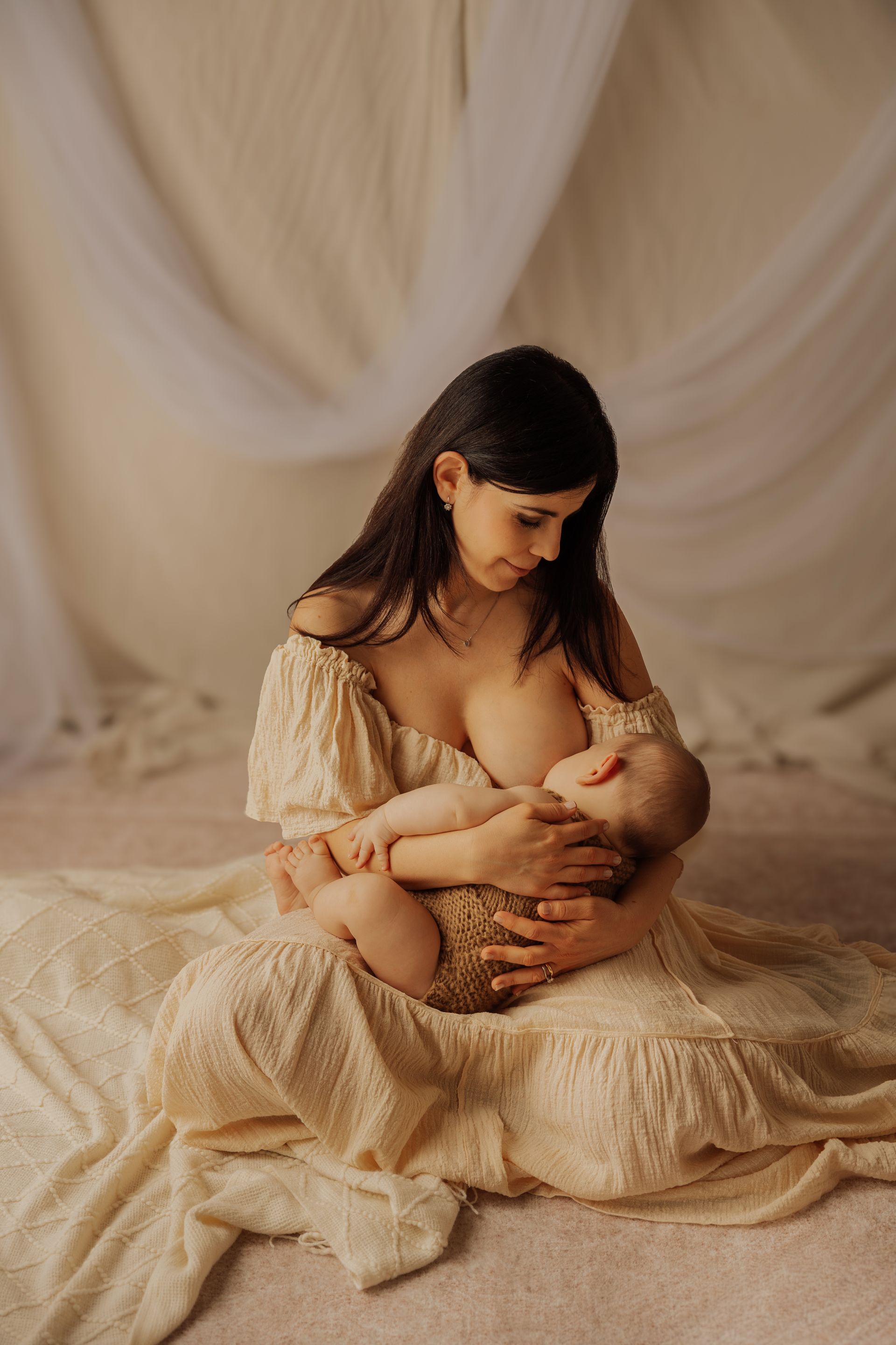 Una mamma con abito avorio allatta il suo bambino seduta a terra davanti a tende chiare