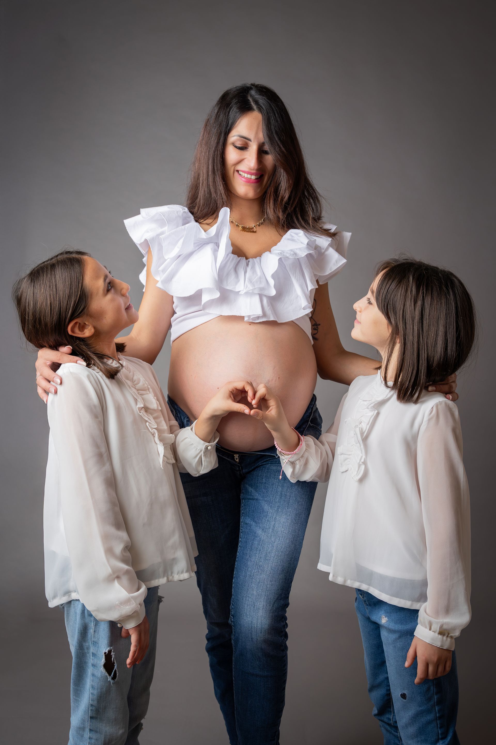 Due sorelle in camicie bianche abbracciano la mamma incinta con il pancione scoperto, fondo grigio