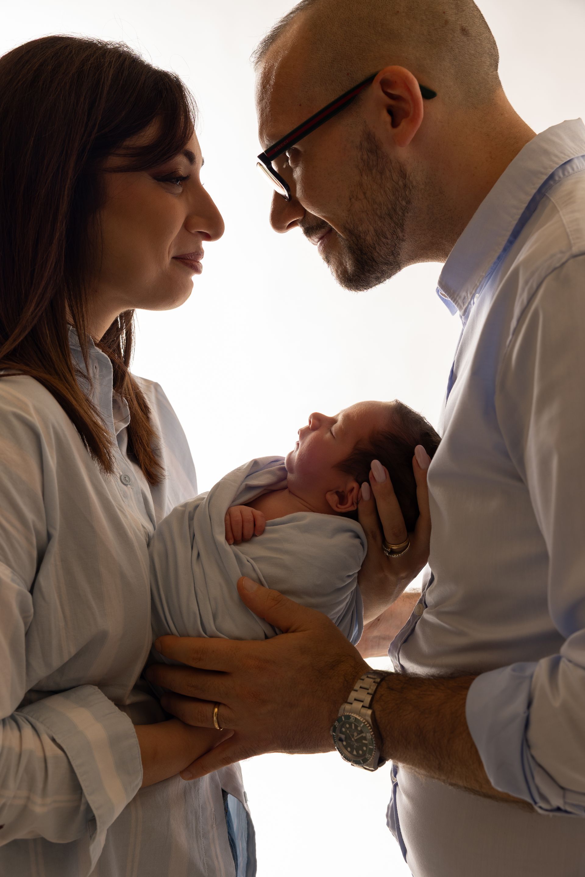 Mamma e papà si guardano mentre tengono insieme il neonato fasciato, in controluce su fondo bianco