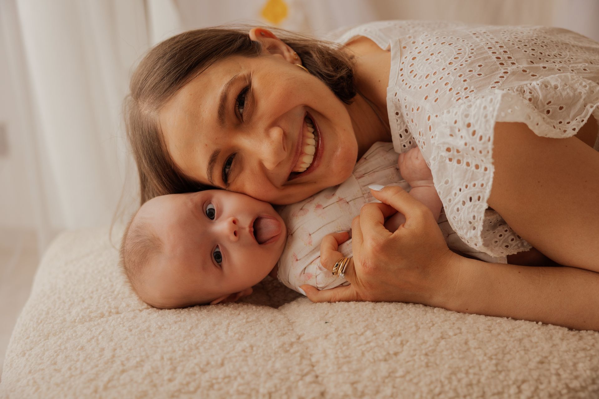 Mamma sorridente appoggia la guancia a quella della figlia neonata distesa su un tappeto bianco morbido