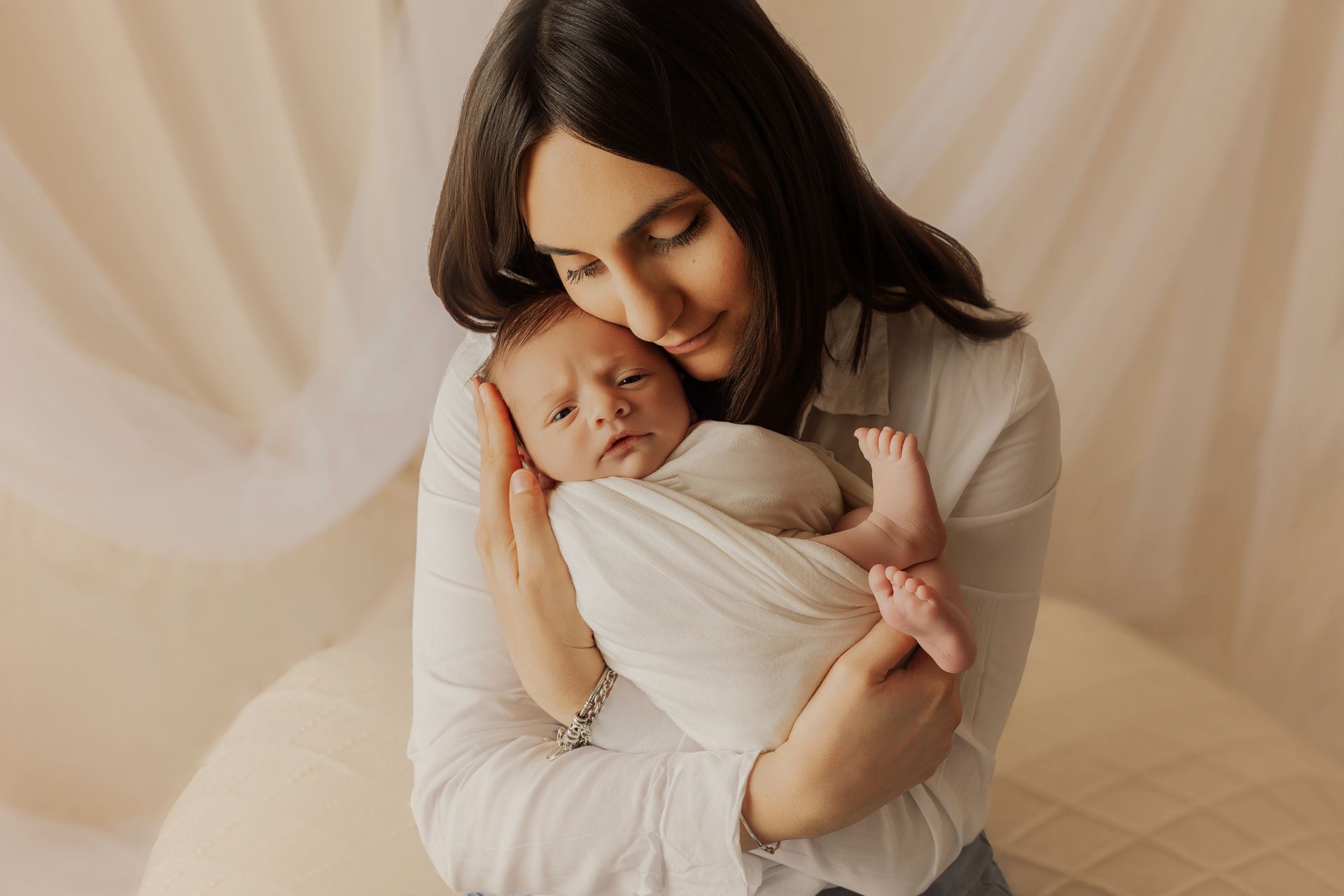 Mamma in camicia bianca stringe al petto il neonato fasciato e lo guarda con tenerezza