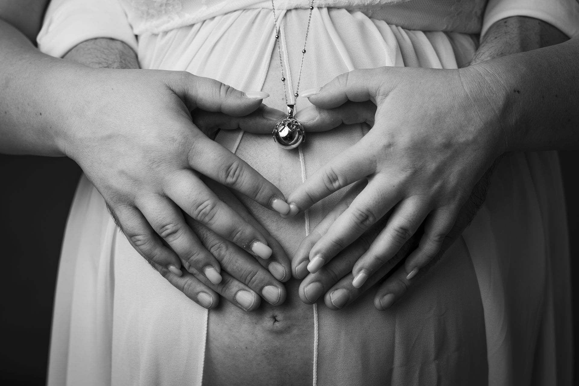 Le mani della coppia formano un cuore sul pancione attorno a un ciondolo, in monocromia