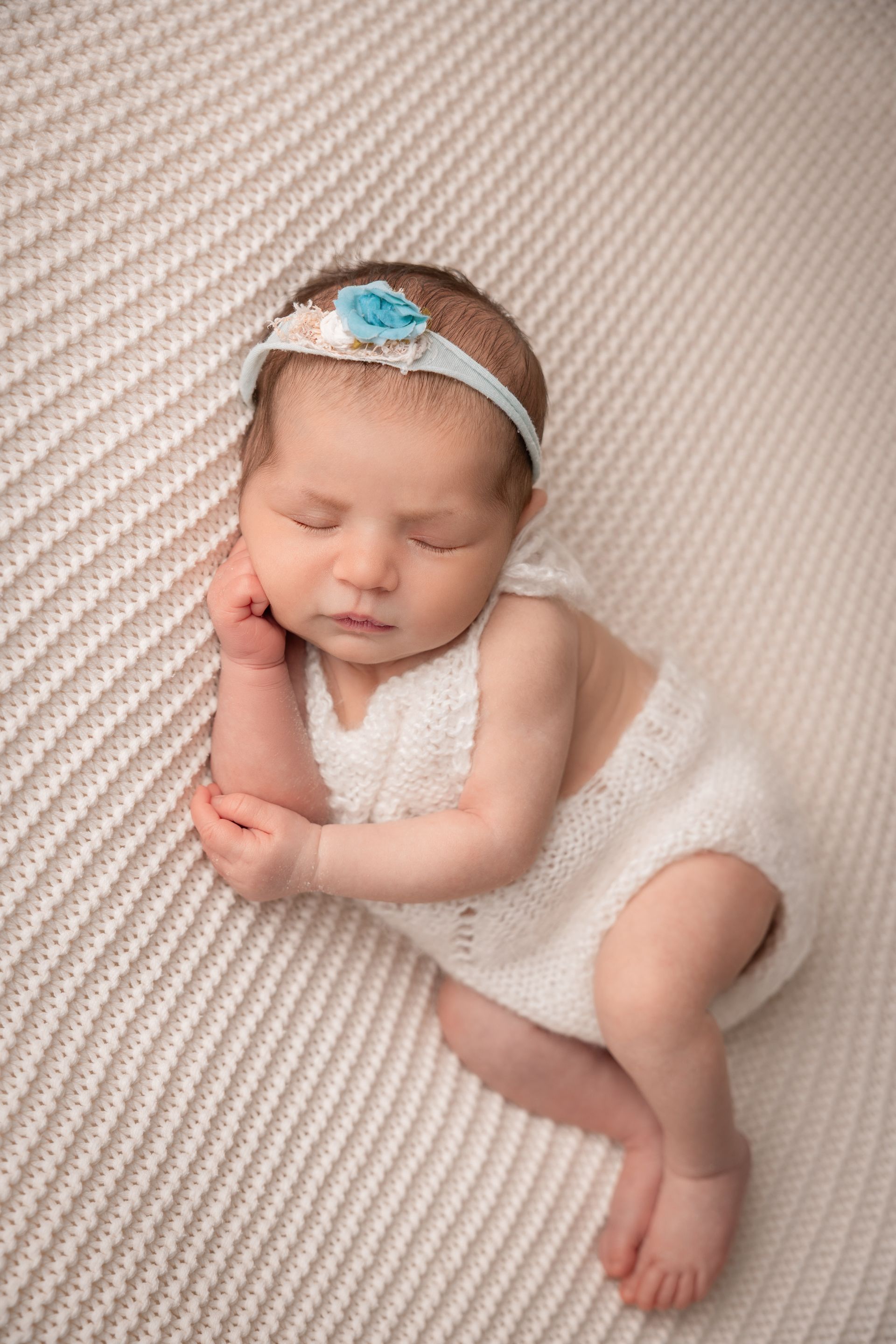Neonata con fascia e fiorellino azzurro in tutina bianca a maglia, dorme raccolta su un fondo panna