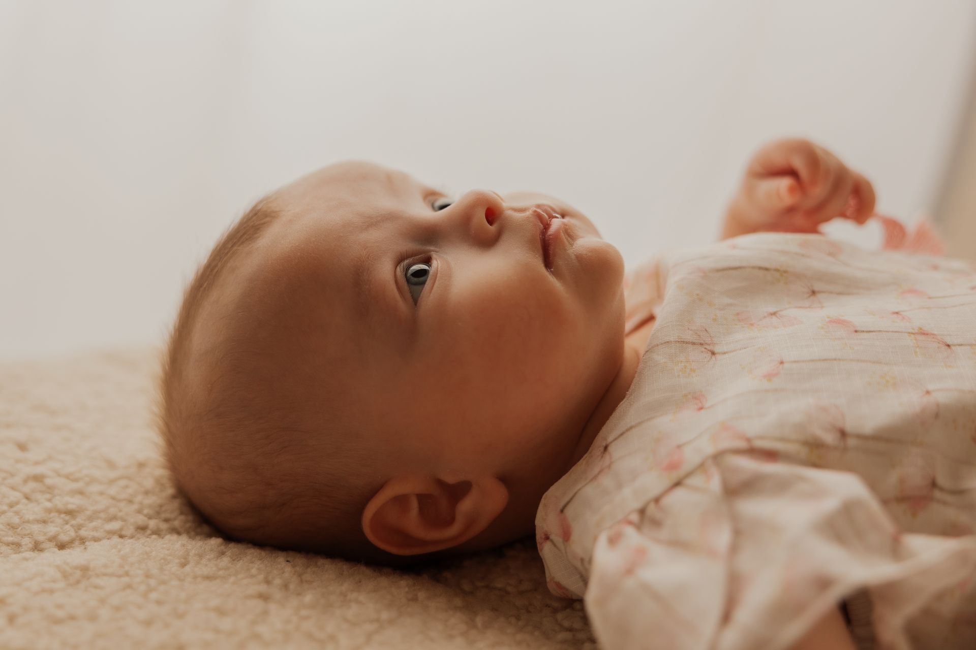 Neonata di profilo sdraiata su una coperta morbida beige, bocca socchiusa e abitino rosa chiaro