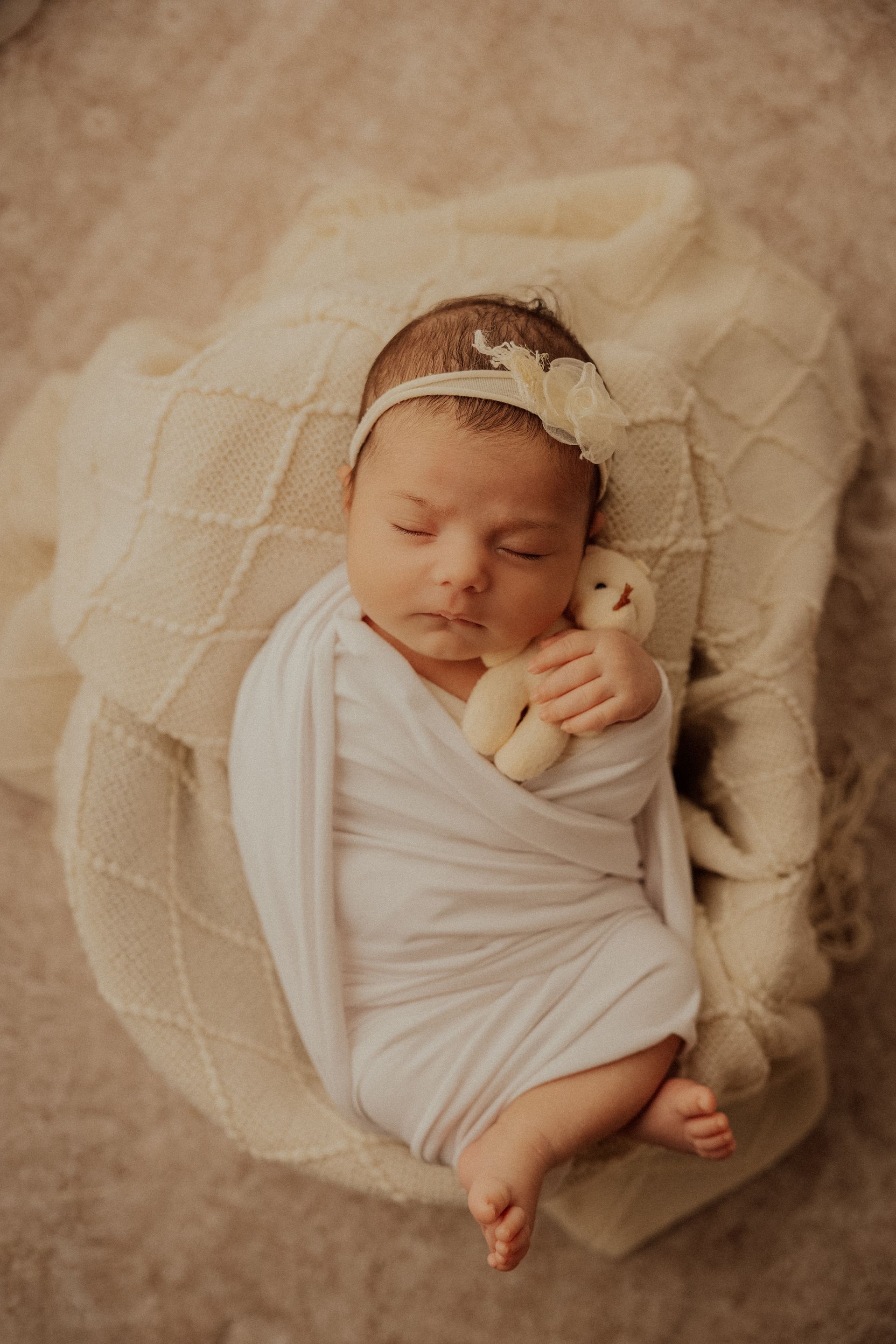 Neonata con fiore bianco tra i capelli dorme fasciata su coperta crema accanto a un peluche