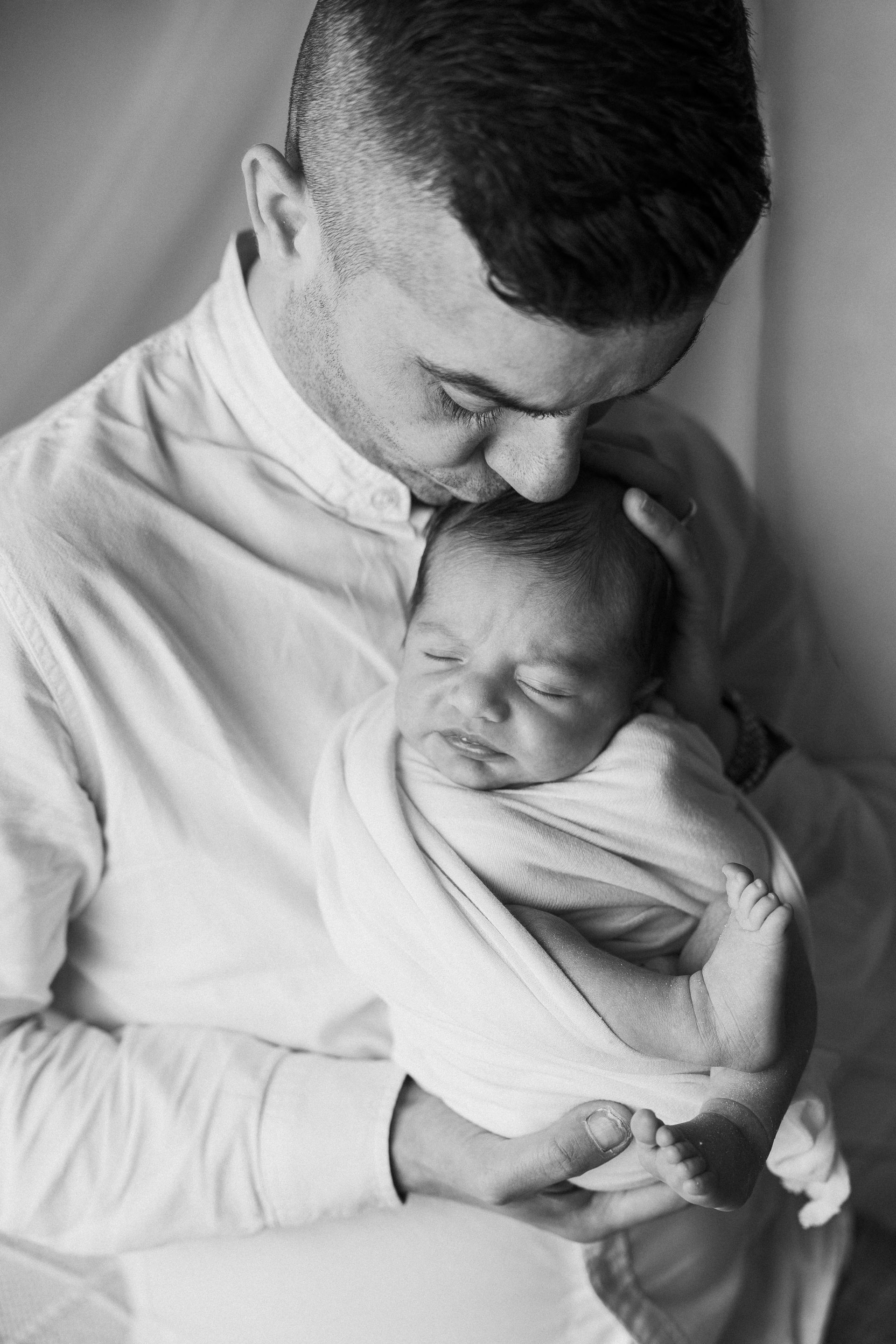 Papà culla il neonato fasciato fra le braccia e gli sfiora la testa con un bacio, scatto monocromatico