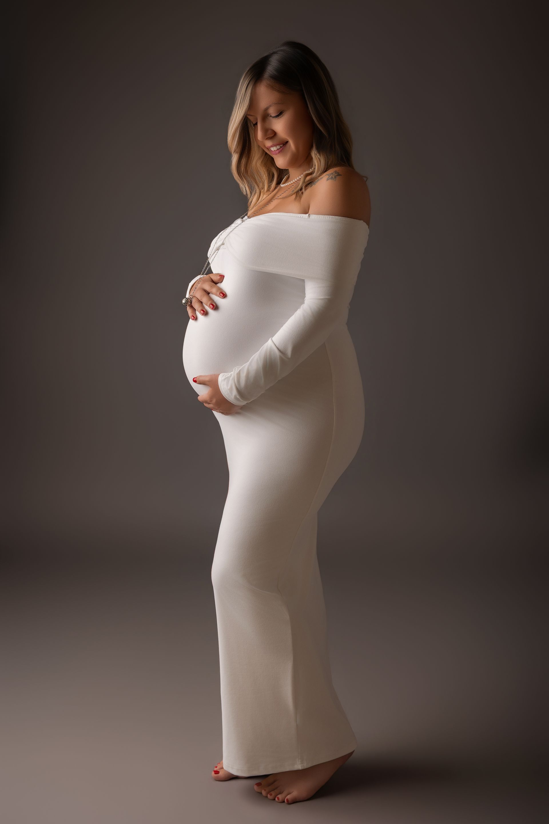 Futura mamma di profilo in lungo abito bianco spalle scoperte, mani sul ventre su fondo bruno