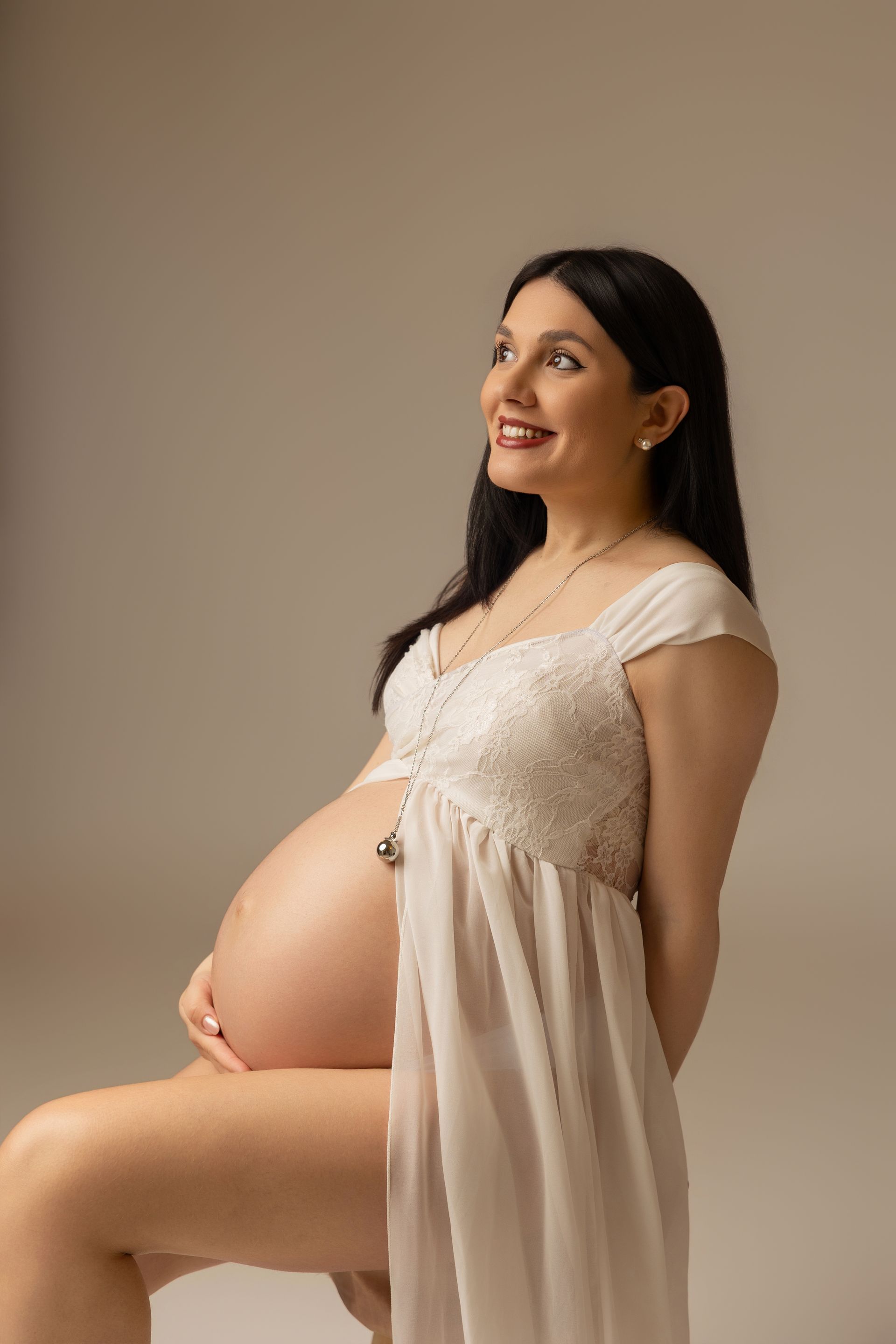 Futura mamma seduta in abito di pizzo panna con il ventre scoperto, sorride guardando di lato