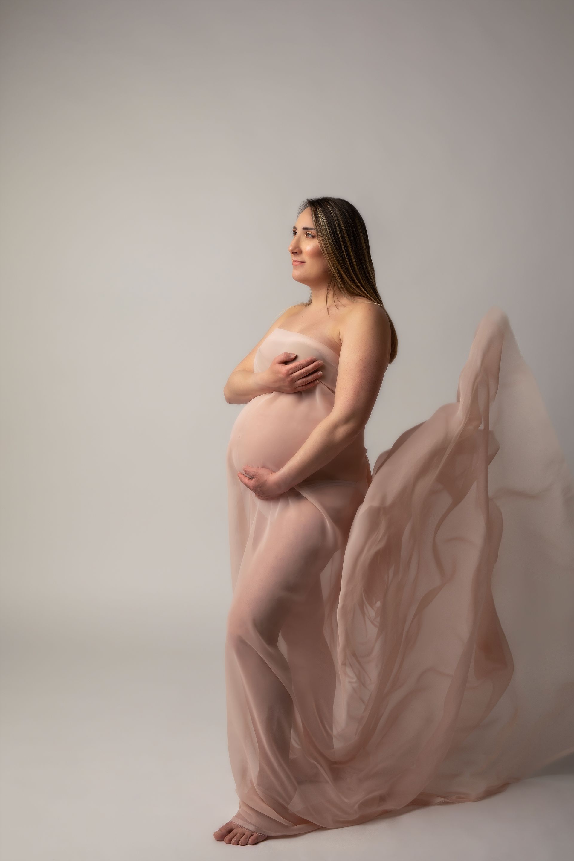 Futura mamma di profilo con pancione scoperto avvolta in un velo rosa cipria su fondo beige
