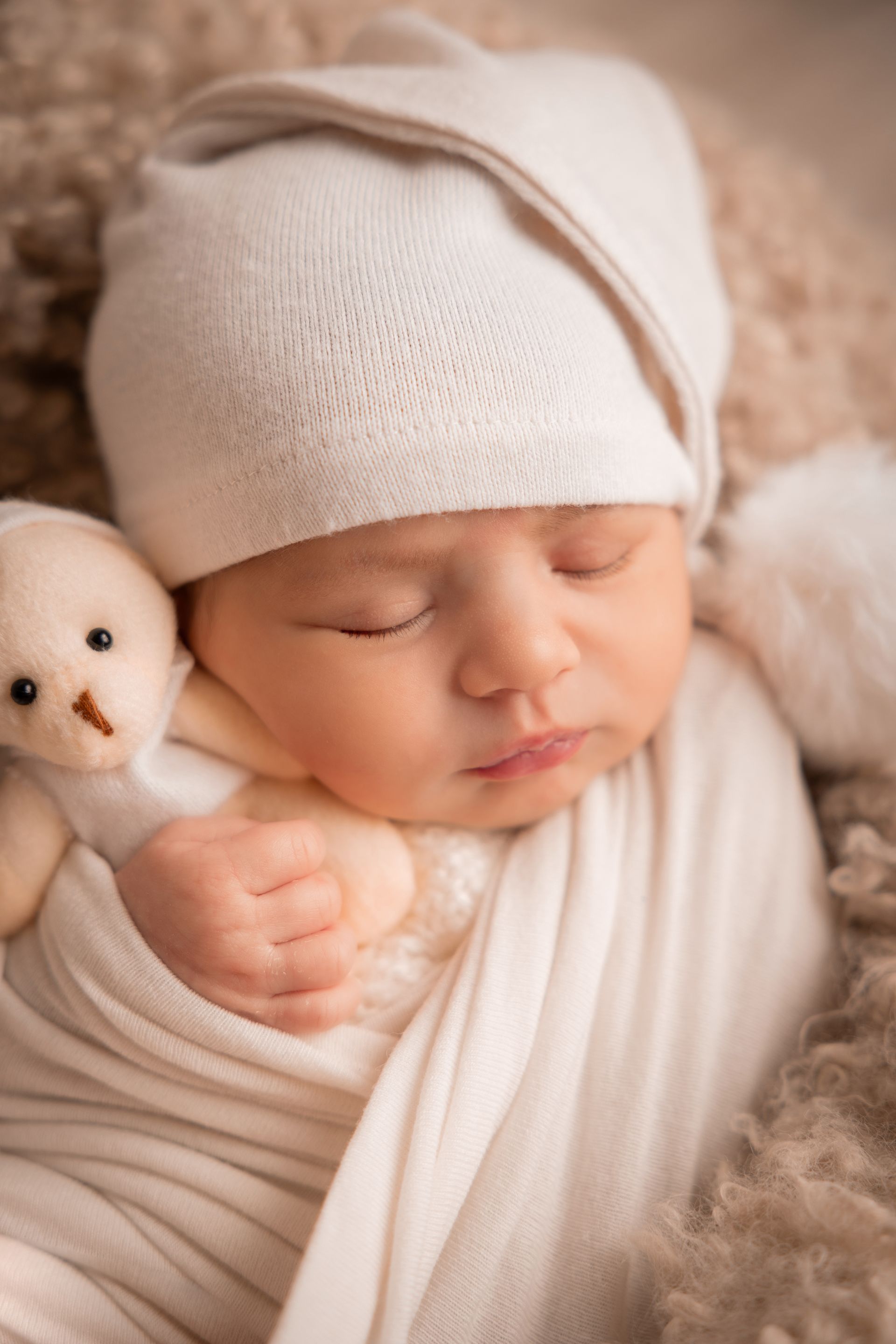 Primo piano di un neonato addormentato fasciato di bianco con cuffietta chiara accanto a un orsetto
