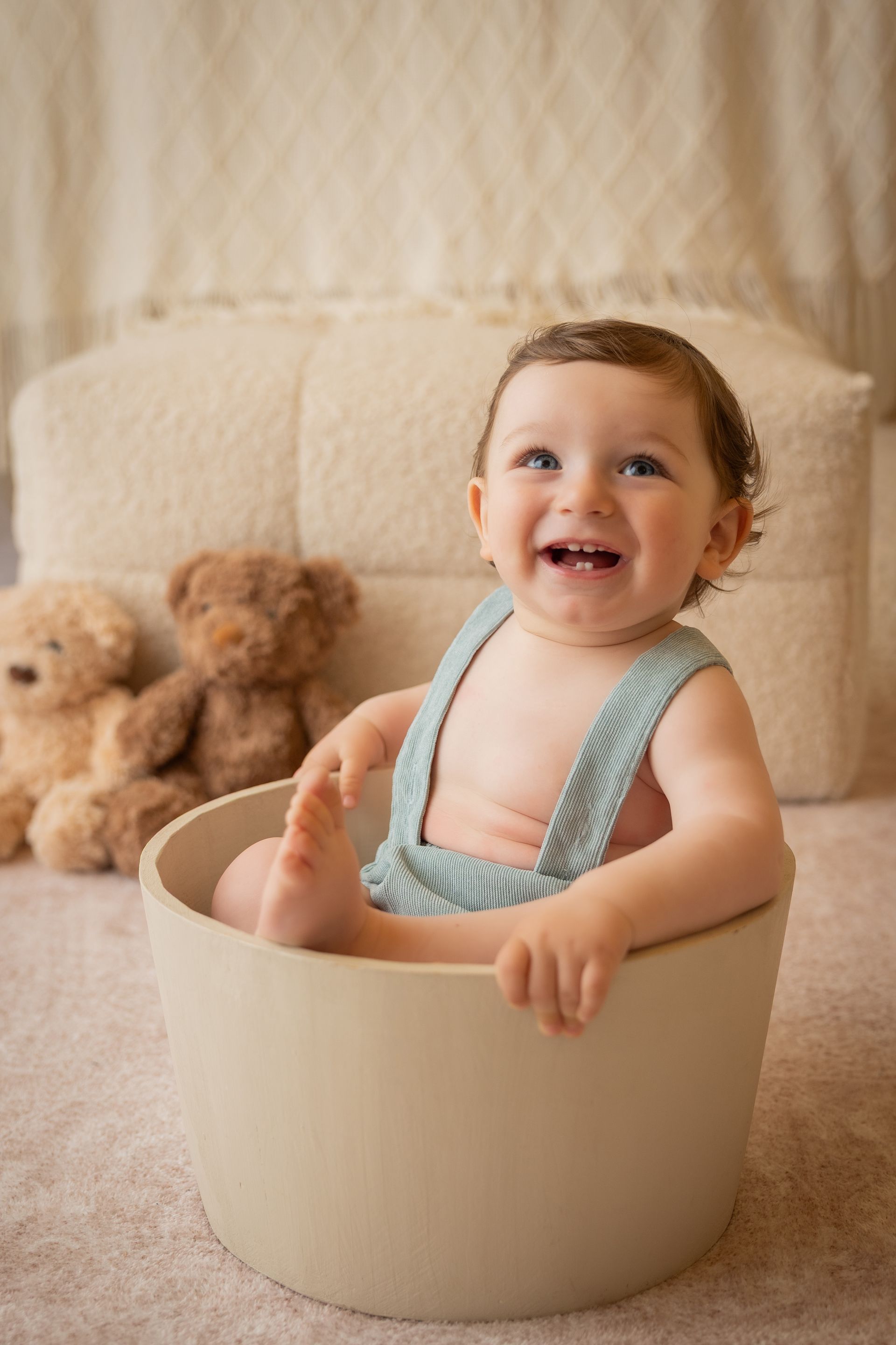 Bambino in salopette verde salvia ride seduto in un mastello, due orsetti appoggiati alle sue spalle