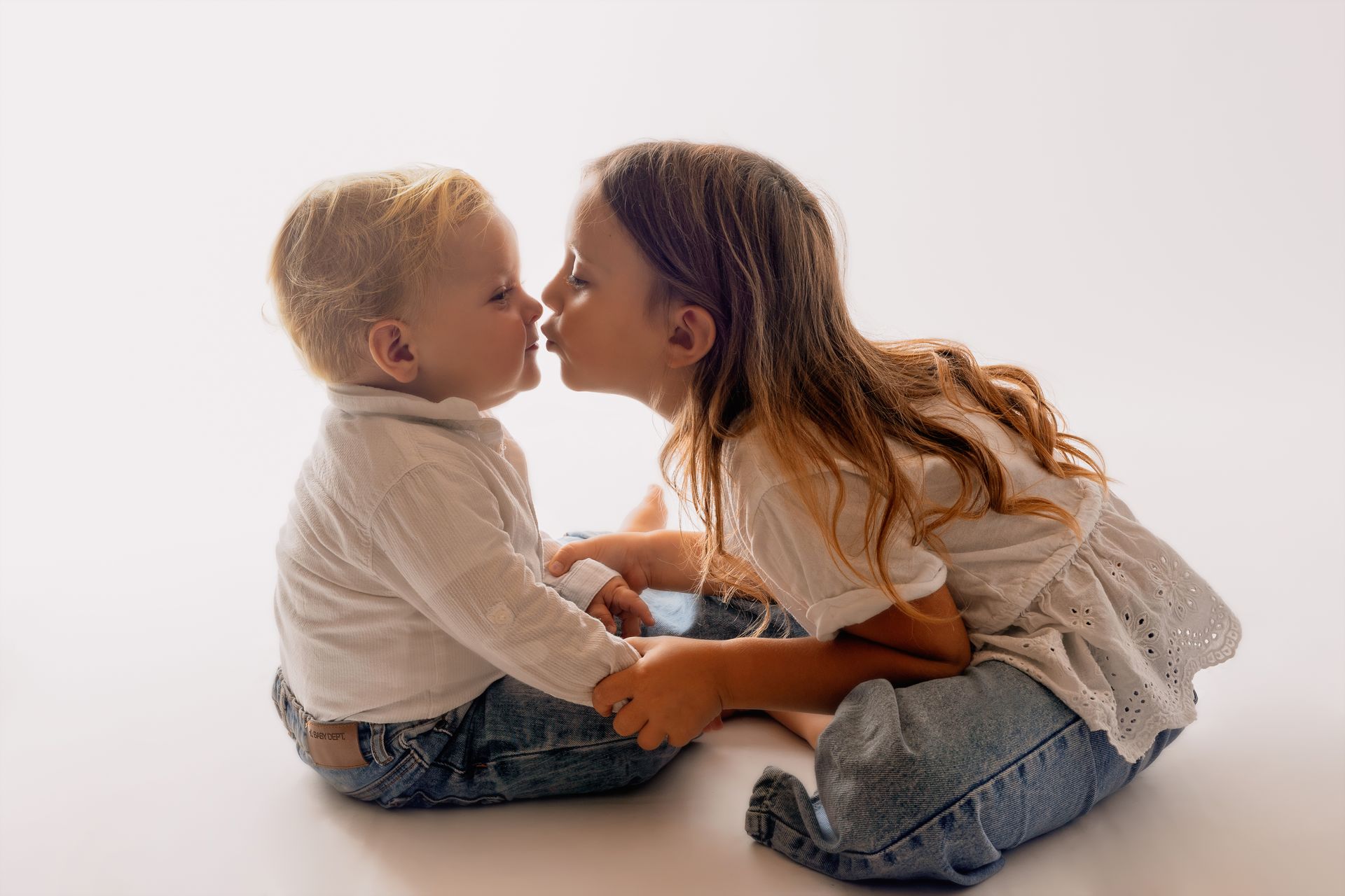Bambina più grande bacia il fratellino sulle labbra, entrambi in camicia bianca e jeans su fondo chiaro